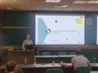 Előadó teremben az előadó prezentációt vetít ki. Nézőtéren a hallgatóságból is látszódik 2 ember.